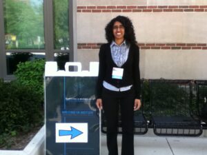 Tamil woman with long curly black hair stands next to a blue folding sign for Computers & Writing 2011, in front of green hedges and a brick and stone building.
