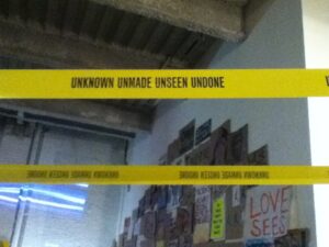 Yellow caution tape reading "Unknown Unmade Unseen Undone" strung across a doorway at the "Occupy Wall Street: No Comment" gallery exhibit.
