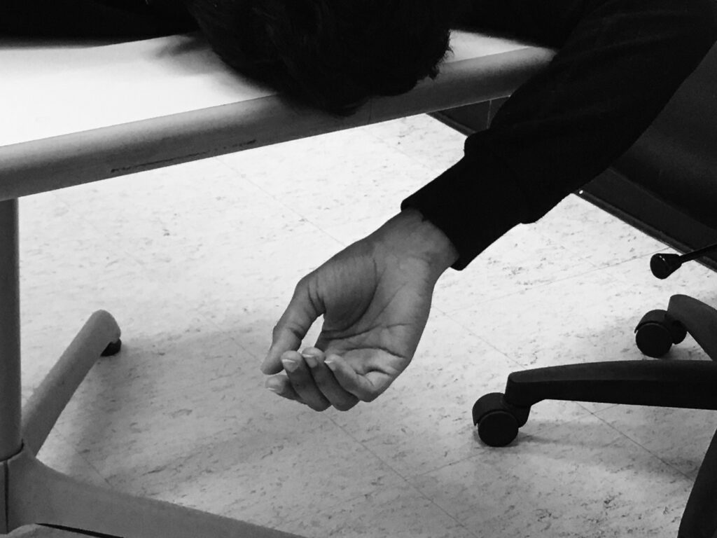 Black and white photo of a limp hand hanging off the edge of a desk with the wheels of rolling chairs visible
