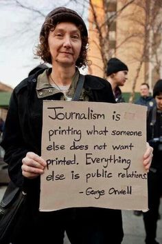 A white woman in a jacket holds a cardboard sign: Journalism is printing what someone else does not want printed. Everything else is public relations. - George Orwell