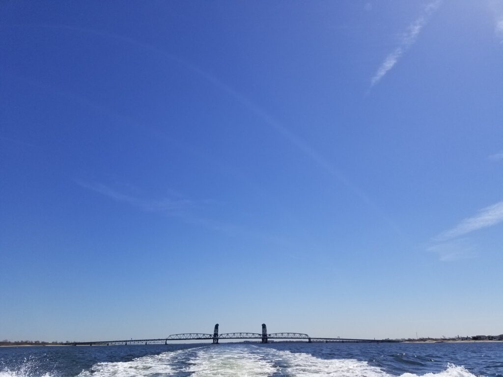 The whitewater wake behind a boat off of Coney Island shore, a bridge visible in the distance, blue skies overhead.