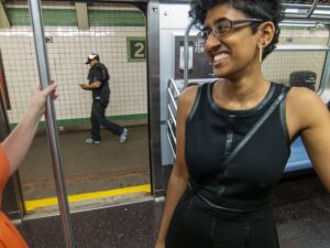 Tamil woman in a black skater dress with leather trim stands smiling in a subway car with open doors.