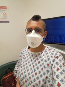 Tamil woman in a hospital gown with black faux hawk, green glasses, and white N95 mask sits in front of a wall-mounted monitor.