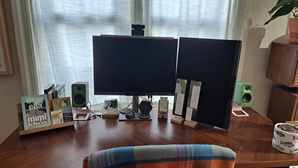 A clean desktop with dual monitors in front of double windows, with two memo stands, speakers, paperweights, and a clock.