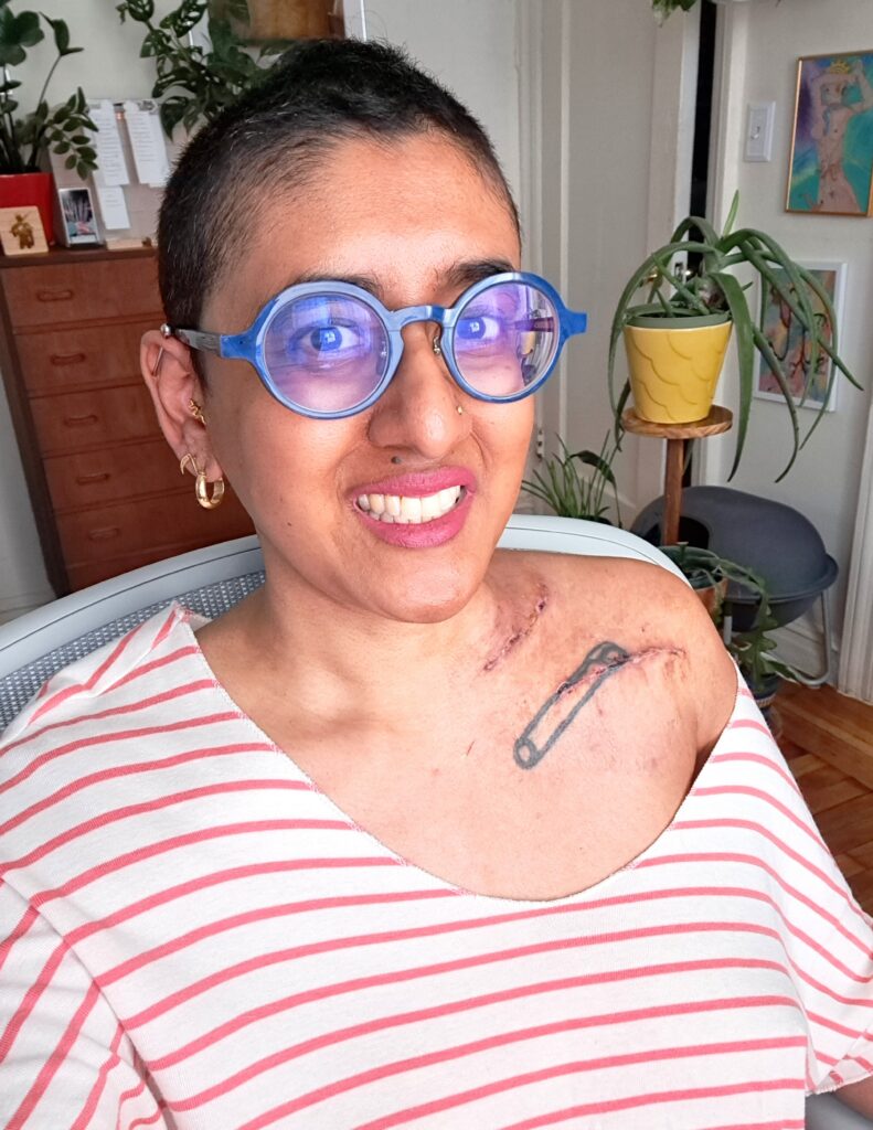 Tamil woman wearing blue glasses, a pink-and-white striped shirt, and pink lipstick sitting in an office chair with houseplants and a dresser in the background.