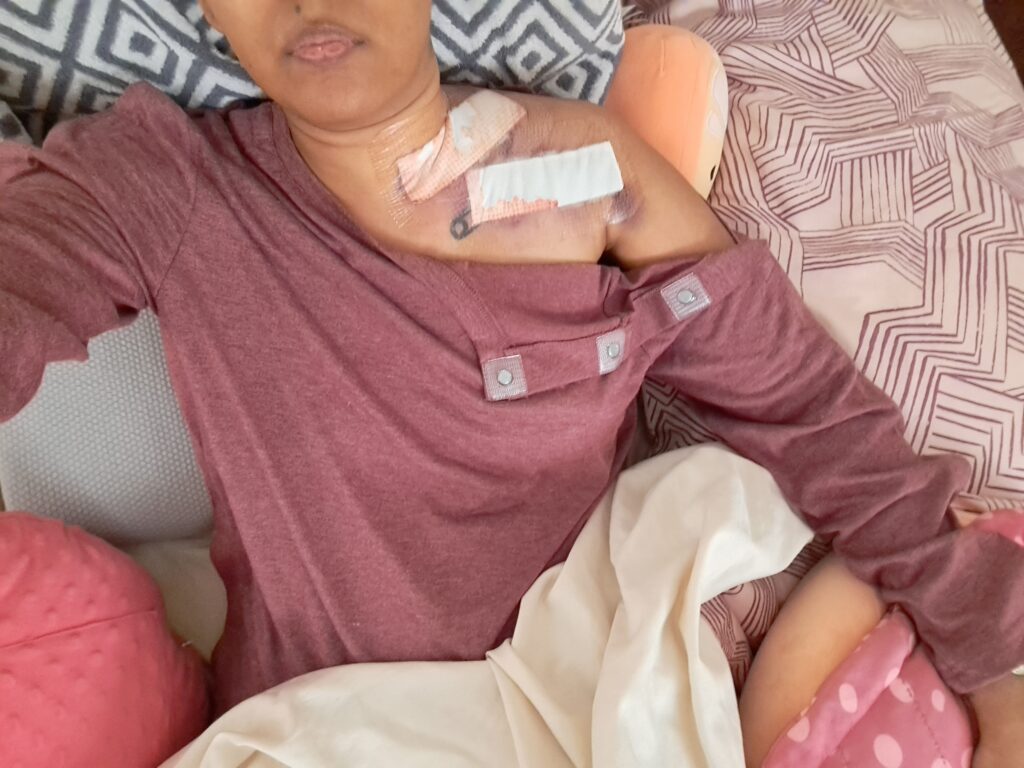 Tamil woman on a reclining cushion in bed with bloody bandages on her neck and Squishmallows around her.